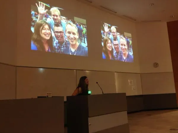 Presenter at a podium beside two projector screens showing forum attendees waving